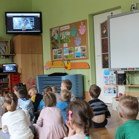 Fotografia kolorowa. Dzieci słuchają i oglądają bibliotekarkę, która czyta im bajkę i wyświetla się na telewizorze. 