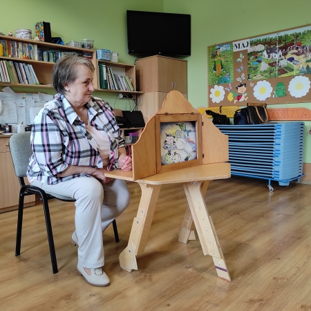 Fotografia kolorowa. Bibliotekarka przedstawia dzieciom historię za pomocą drewnianego teatrzyku. 