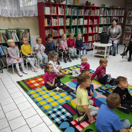 Fotografia kolorowa. Grupa przedszkolaków siedzi na dywanie w bibliotece. 