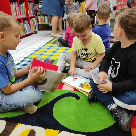 Fotografia kolorowa. Grupa przedszkolaków siedzi na dywanie w bibliotece i ogląda książki. 