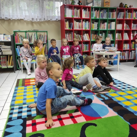Fotografia kolorowa. Grupa przedszkolaków siedzi na dywanie w bibliotece. 