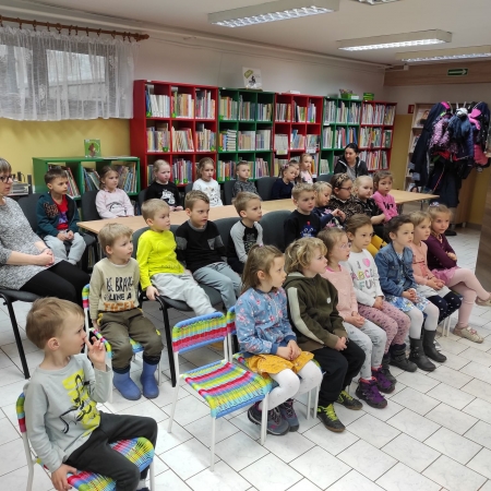 Fotografia kolorowa. Grupa dzieci siedzi na krzesłach i przysłuchuje się bibliotekarce.