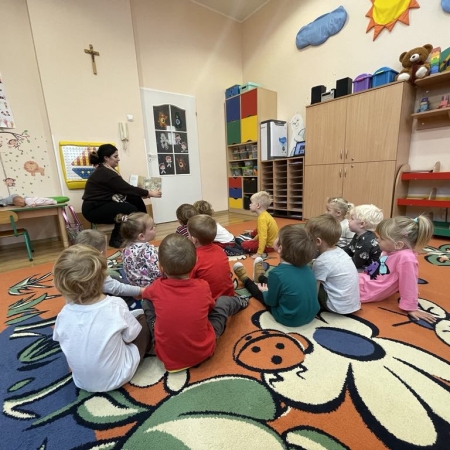 Fotografia kolorowa. Zdjęcie grupowe w przedszkolu. Dzieci słuchają opowiadania, które czyta im bibliotekarka. 