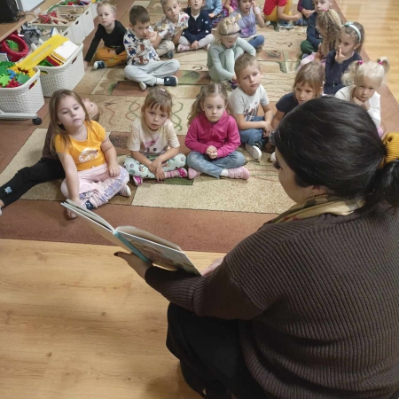 Fotografia kolorowa. Zdjęcie grupowe w przedszkolu. Dzieci słuchają opowiadania, które czyta im bibliotekarka. 