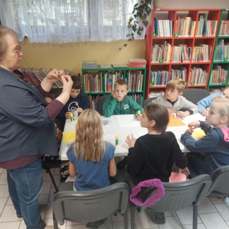 Fotografia kolorowa. Zdjęcie grupowe w bibliotece. Bibliotekarka wyjaśnia jak wykonać prace plastyczne. 