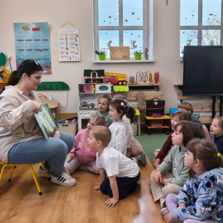 Fotografia kolorowa. Grupa przedszkolaków siedzi na dywanie i słucha opowieści czytanych przez bibliotekarkę.