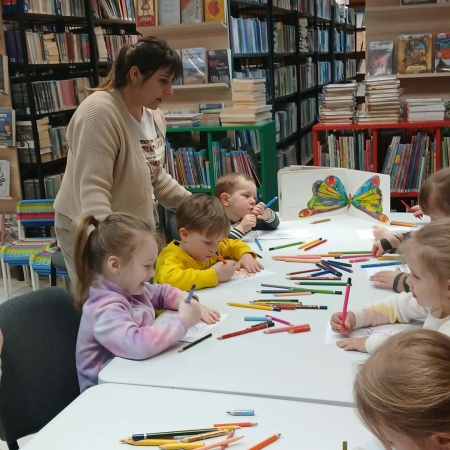 Fotografia kolorowa. Grupa dzieci siedzi przy stole w bibliotece i wykonuje prace plastyczne.