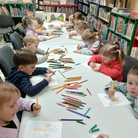 Fotografia kolorowa. Grupa dzieci siedzi przy stole w bibliotece i wykonuje prace plastyczne.