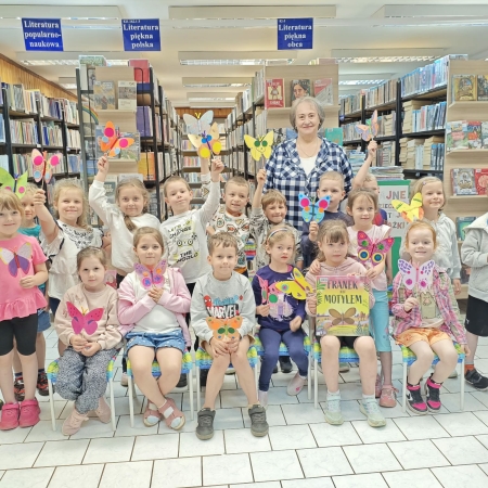 Fotografia kolorowa. Grupa przedszkolaków we wnętrzu biblioteki pozuje do zdjęcia grupowego ze swoimi pracami plastycznymi.