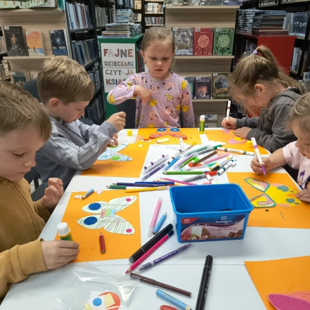 Fotografia kolorowa. Grupa przedszkolaków we wnętrzu biblioteki wykonuje prace plastyczne.