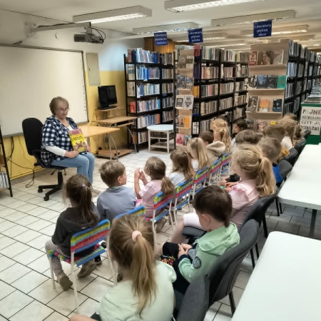 Fotografia kolorowa. Grupa przedszkolaków siedzi na krzesełkach we wnętrzu biblioteki i słucha opowieści. 