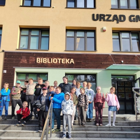 Fotografia kolorowa. Dzieci i bibliotekarka przed budynkiem biblioteki. 